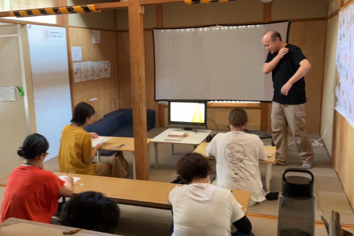 須坂市の放課後等デイサービス「さくらぼ須坂中央教室」にて、「自分らしく歩むために」をテーマに講演するMDMリハ整体院の作業療法士。会場のスクリーンには「先回りをしない見守り方と、頼れる場所の作り方」の文字が映されている